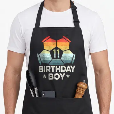 Player 11th birthday apron - Rainbow soccer ball with number 11 and birthday boy text flanked by stars — shown on a black cotton canvas apron against a white background