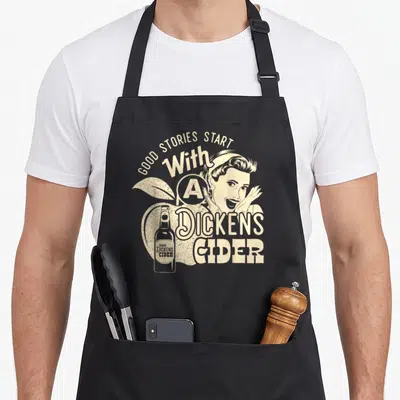 Hard apple apron - Beige retro woman and bottle over apple with arched text Good Stories Start With A Dickens Cider — shown on a black cotton canvas apron against a white background