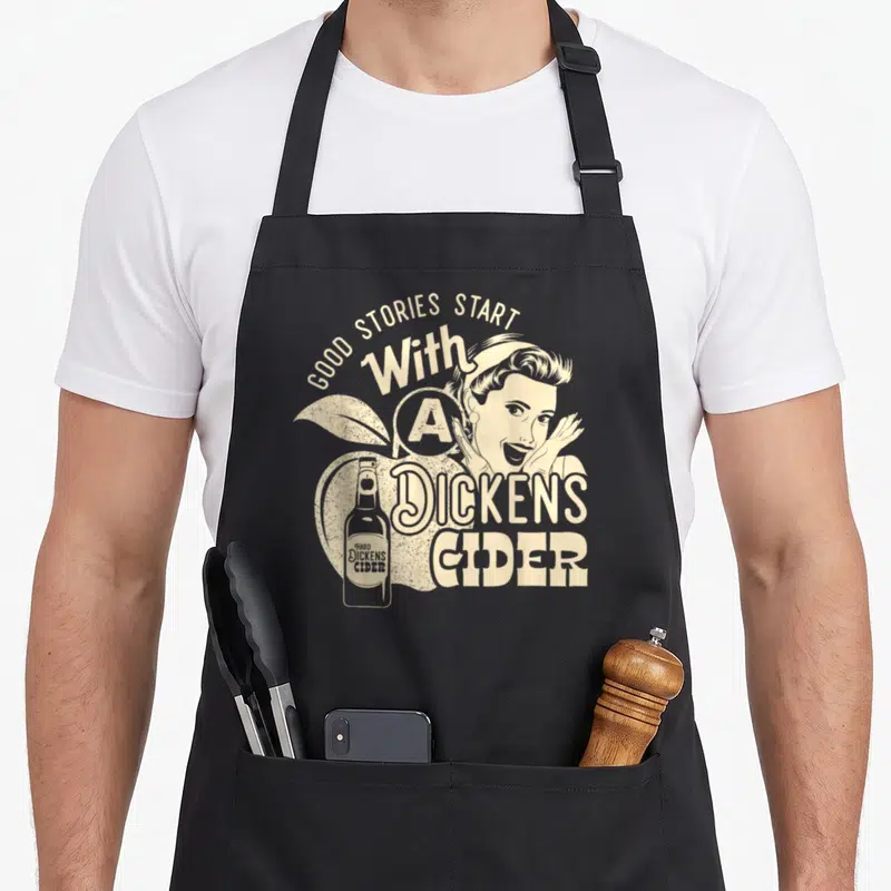 Hard apple apron - Beige retro woman and bottle over apple with arched text Good Stories Start With A Dickens Cider — shown on a black cotton canvas apron against a white background