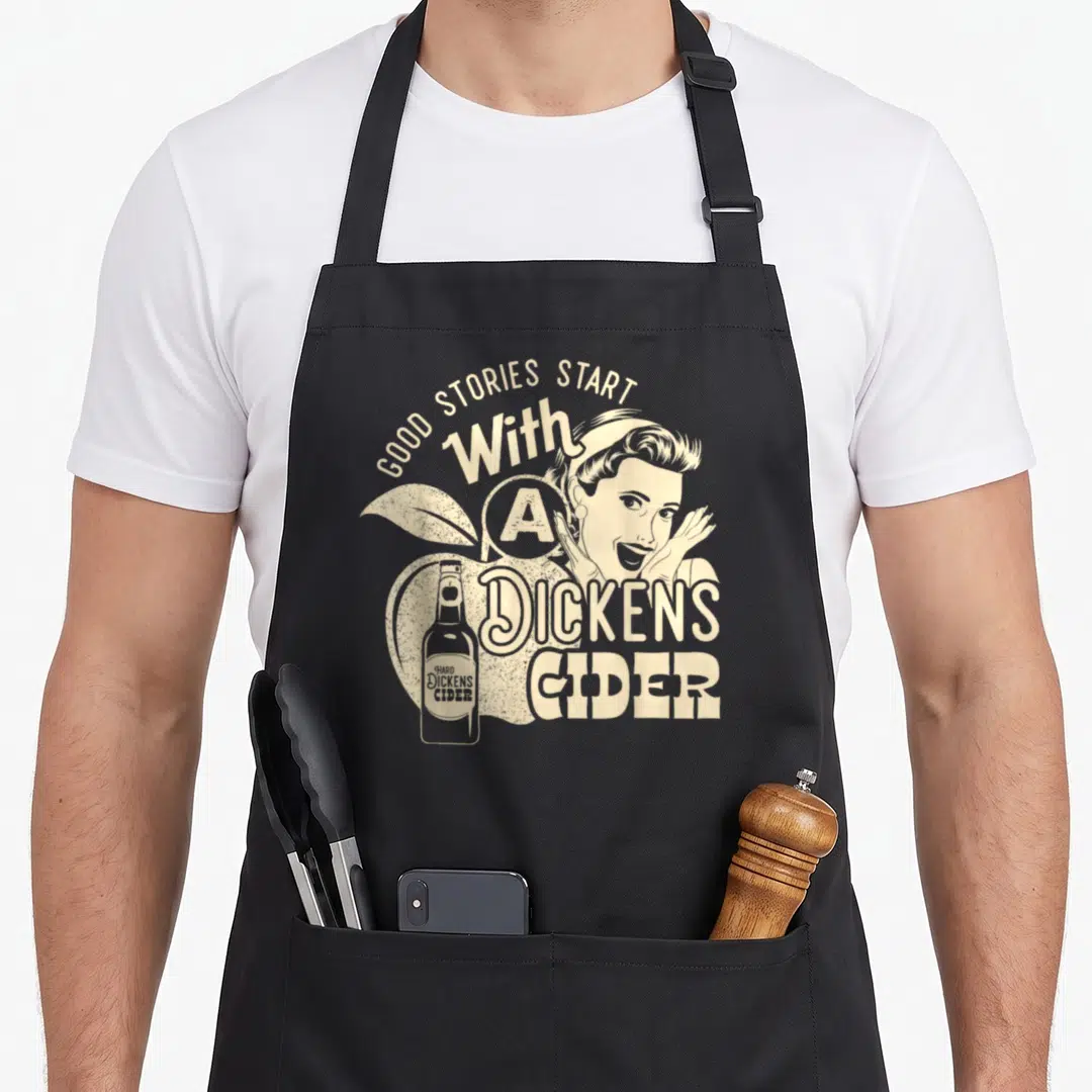 Hard apple apron - Beige retro woman and bottle over apple with arched text Good Stories Start With A Dickens Cider — shown on a black cotton canvas apron against a white background