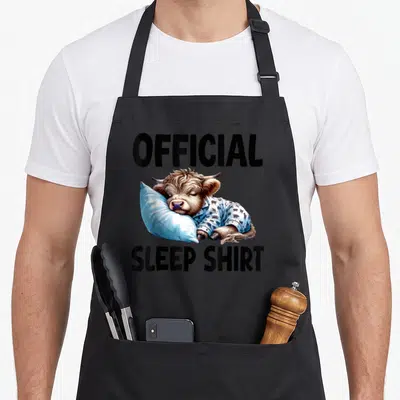 Sleepshirt highland cow apron - Realistic brown cow lying on blue pillow wearing pajamas with official sleep shirt text — shown on a black cotton canvas apron against a white background