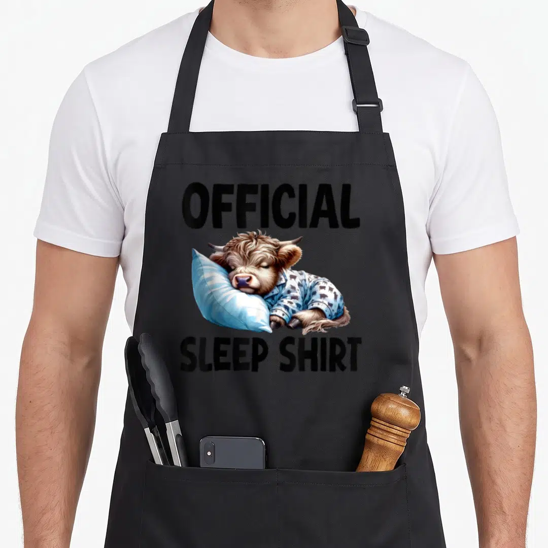 Sleepshirt highland cow apron - Realistic brown cow lying on blue pillow wearing pajamas with official sleep shirt text — shown on a black cotton canvas apron against a white background
