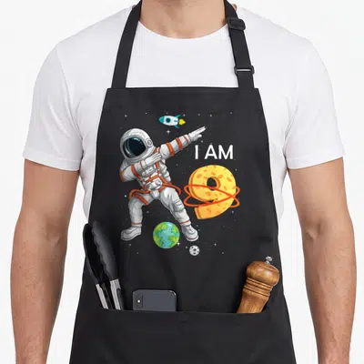 Space birthday apron - Dabbing astronaut next to I AM text and cheese-textured number 9 with rocket and planets — shown on a black cotton canvas apron against a white background