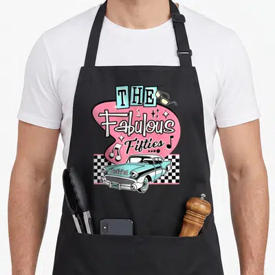 50s sock rockabilly apron - The Fabulous Fifties 1958 text above light blue car on checkered strip with pink shape — shown on a black cotton canvas apron against a white background