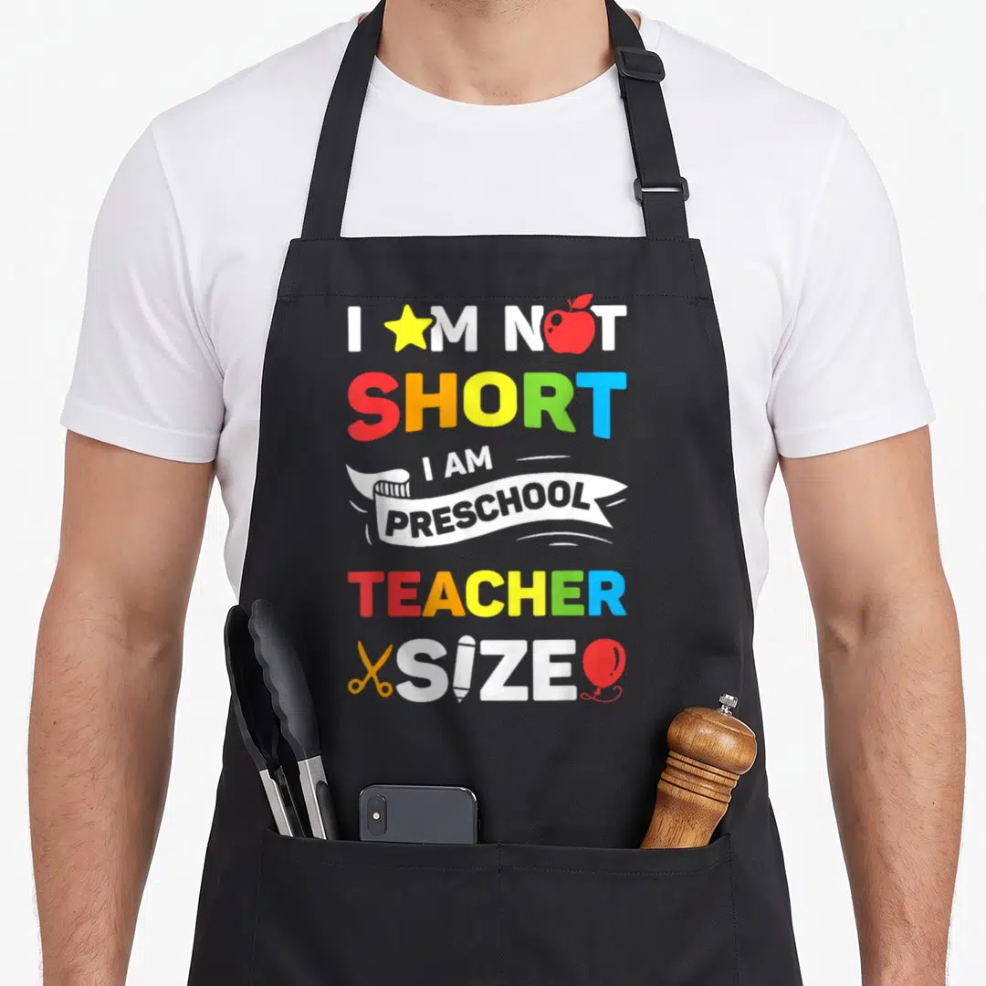 Funny preschool teacher apron - Colorful stacked text with star apple scissors pencil balloon replacing letters — shown on a black cotton canvas apron against a white background