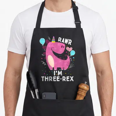Rawr three t rex apron - Pink dinosaur with stars wearing a party hat surrounded by blue and pink balloons and confetti — shown on a black cotton canvas apron against a white background