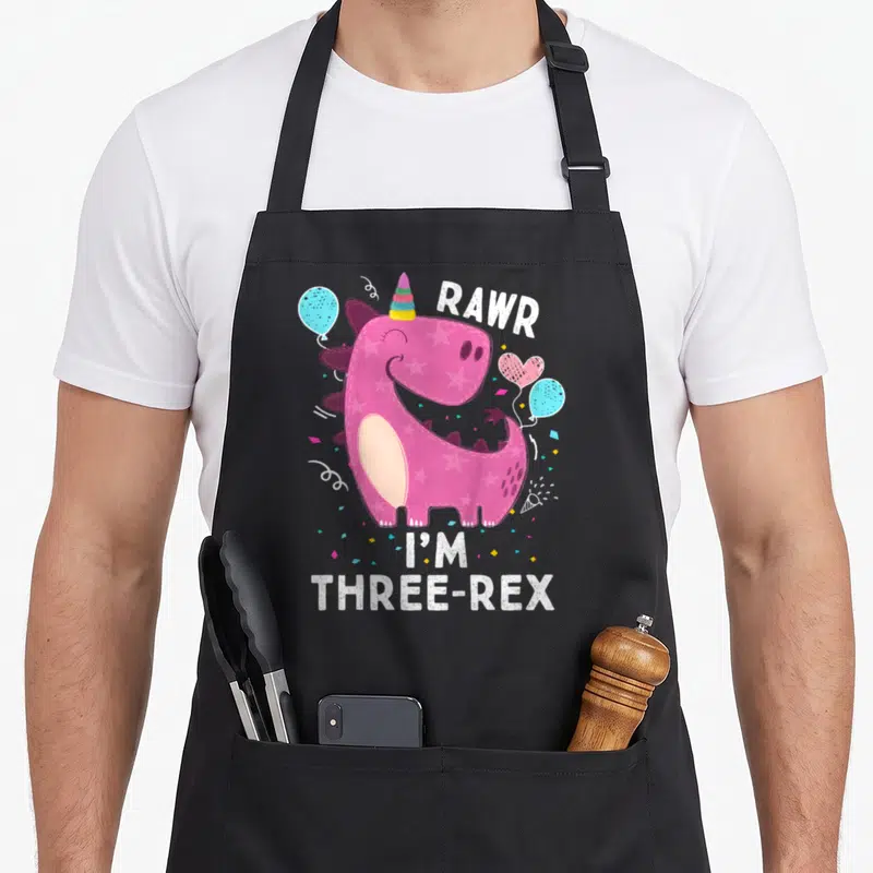 Rawr three t rex apron - Pink dinosaur with stars wearing a party hat surrounded by blue and pink balloons and confetti — shown on a black cotton canvas apron against a white background
