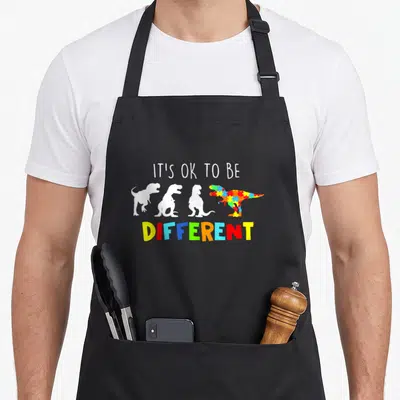 Awareness dinosaur apron - IT'S OK TO BE DIFFERENT text above four dinosaur silhouettes including one with colorful — shown on a black cotton canvas apron against a white background