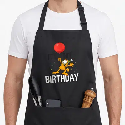 Celebration birthday apron - Orange Garfield holding red balloon with IT'S MY BIRTHDAY text and confetti — shown on a black cotton canvas apron against a white background