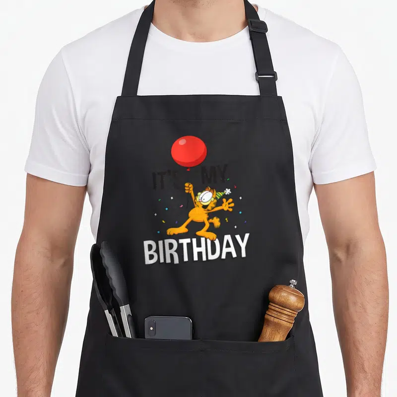 Celebration birthday apron - Orange Garfield holding red balloon with IT'S MY BIRTHDAY text and confetti — shown on a black cotton canvas apron against a white background