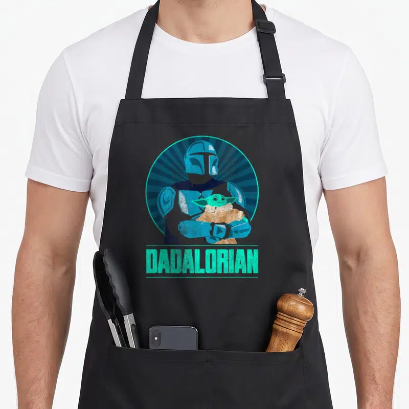 Funny dadalorian father day apron - Cartoon Mandalorian holding Grogu in blue and tan circle above teal Dadalorian text — shown on a black cotton canvas apron against a white background