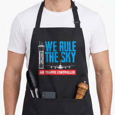 Rule sky apron - We Rule The Sky text with tower plane and Air Traffic Controller banner in blue white red — shown on a black cotton canvas apron against a white background
