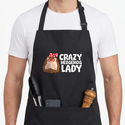 Retro crazy hedgehog lady apron - Cartoon hedgehog with red glasses and bow beside white text CRAZY HEDGEHOG LADY — shown on a black cotton canvas apron against a white background