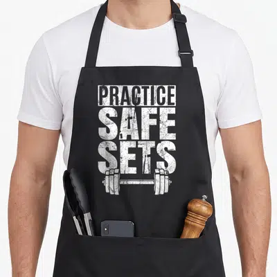 Practice safe apron - White distressed text PRACTICE SAFE SETS above a horizontal dumbbell illustration — shown on a black cotton canvas apron against a white background