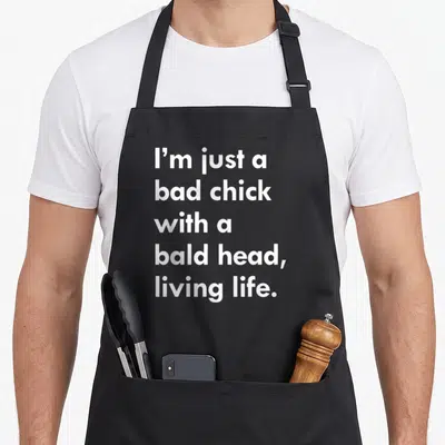 Chick funny bald apron - White text reading I'm just a bad chick with a bald head living life on black fabric — shown on a black cotton canvas apron against a white background