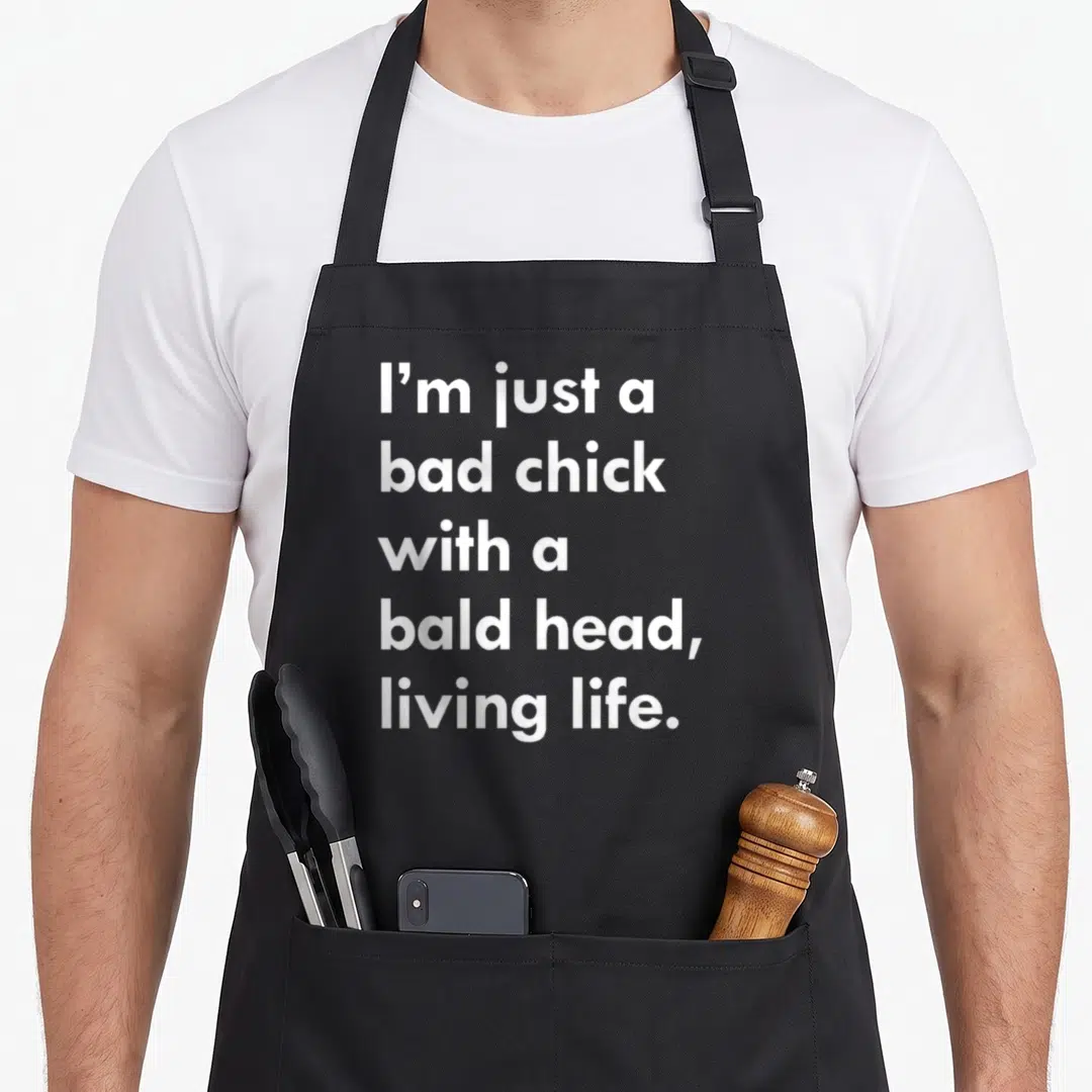 Chick funny bald apron - White text reading I'm just a bad chick with a bald head living life on black fabric — shown on a black cotton canvas apron against a white background