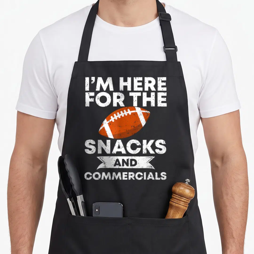 Snack commercial apron - I'M HERE FOR THE SNACKS AND COMMERCIALS text surrounding orange football and ribbon — shown on a black cotton canvas apron against a white background