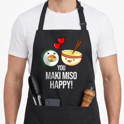 Maki sushi apron - Cartoon maki roll and miso bowl with two hearts and YOU MAKI MISO HAPPY! text — shown on a black cotton canvas apron against a white background