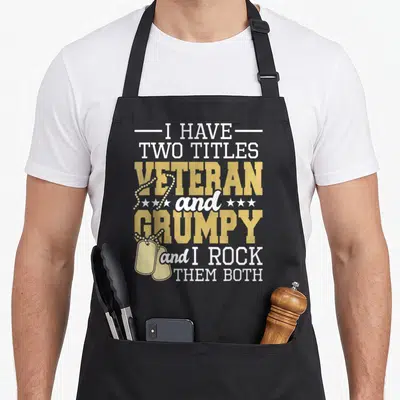 Title patriotic apron - Gold dog tags overlap white and gold stacked text reading I HAVE TWO TITLES VETERAN and GRUMPY — shown on a black cotton canvas apron against a white background