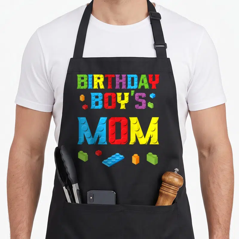 Master builder building aprons - BIRTHDAY BOY'S MOM text formed by blue red yellow green purple building blocks — shown on a black cotton canvas apron against a white background
