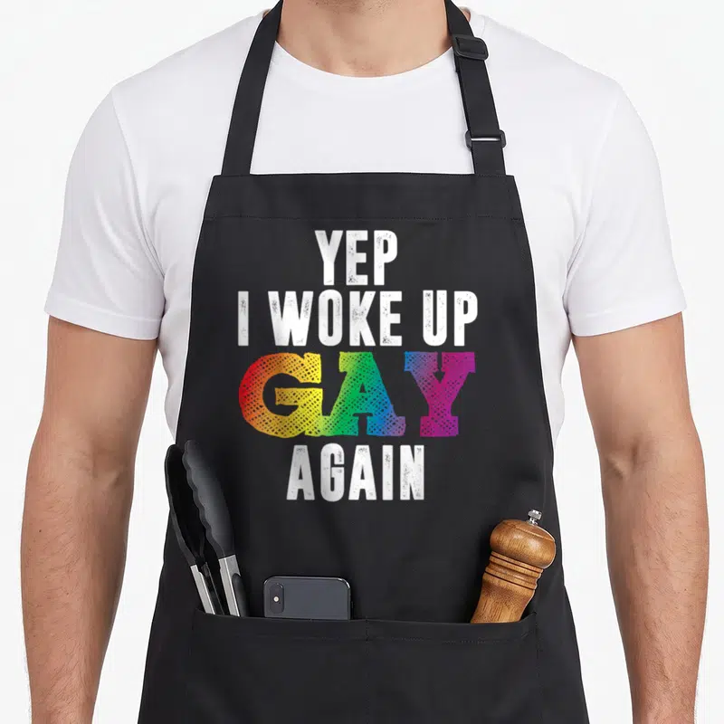 Woke gay pride apron - Stacked white and rainbow gradient text reading YEP I WOKE UP GAY AGAIN — shown on a black cotton canvas apron against a white background