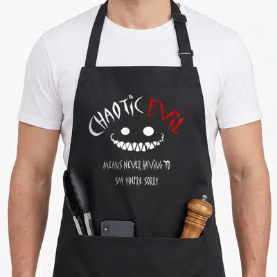 Evil chaotic apron - White and red text Chaotic Evil over a two-eyed monster grin and stacked apology phrase — shown on a black cotton canvas apron against a white background