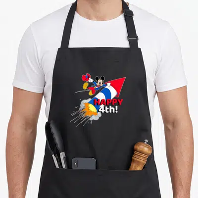 Cute americana fourth of july aprons - Mickey Mouse riding red white blue rocket with HAPPY 4th! text and smoke sparks — shown on a black cotton canvas apron against a white background
