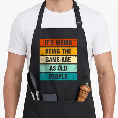 Funny sarcastic weird aprons - Five stacked colored rectangles with distressed grey text reading IT'S WEIRD BEING — shown on a black cotton canvas apron against a white background