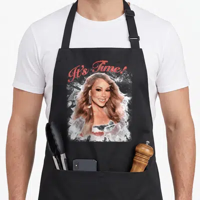 Mariah novelty apron - It's Time! red script above blonde woman breaking white ice shards — shown on a black cotton canvas apron against a white background