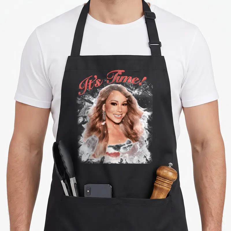 Mariah novelty apron - It's Time! red script above blonde woman breaking white ice shards — shown on a black cotton canvas apron against a white background