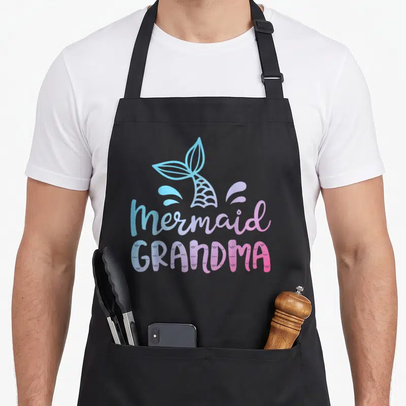 Mermaid grandma in apron - Cartoon mermaid tail and splashes in blue purple pink above stacked text — shown on a black cotton canvas apron against a white background