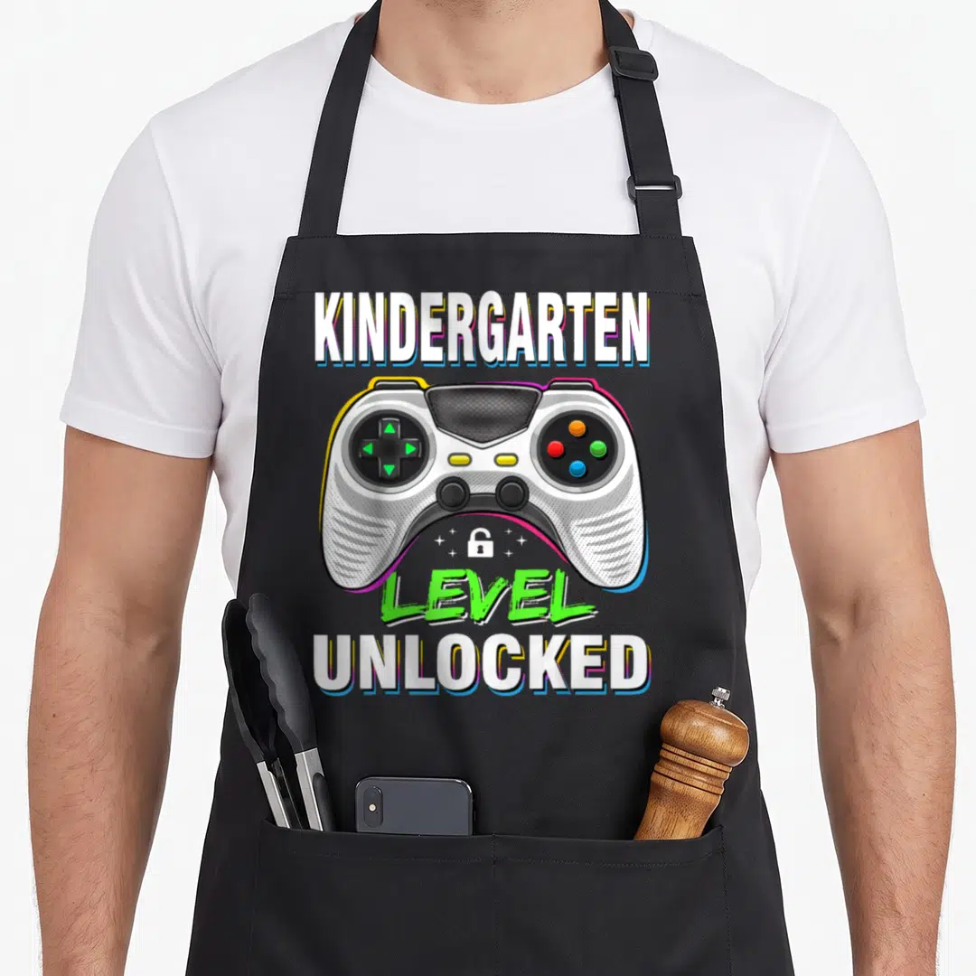 Kindergarten unlocked first apron - Game controller with kindergarten level unlocked text in white grey green blue — shown on a black cotton canvas apron against a white background