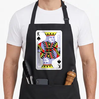 Spade easy apron - King of Spades playing card print with mirrored king figure in white red blue yellow black — shown on a black cotton canvas apron against a white background