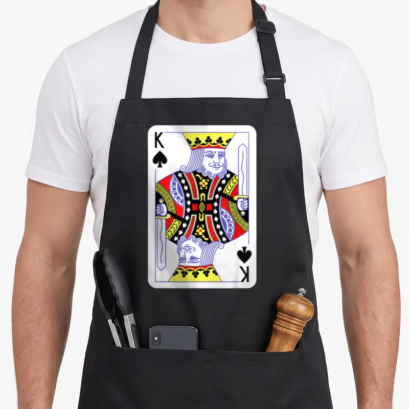 Spade easy apron - King of Spades playing card print with mirrored king figure in white red blue yellow black — shown on a black cotton canvas apron against a white background