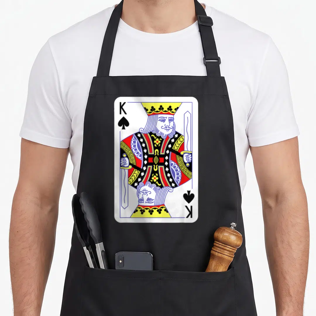 Spade easy apron - King of Spades playing card print with mirrored king figure in white red blue yellow black — shown on a black cotton canvas apron against a white background