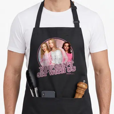 Mean us aprons - Three women in pink circle with text YOU CAN'T SIT WITH US below — shown on a black cotton canvas apron against a white background