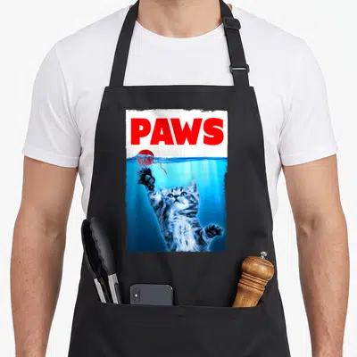 Parody cat apron - PAWS text above underwater kitten reaching for red yarn ball — shown on a black cotton canvas apron against a white background
