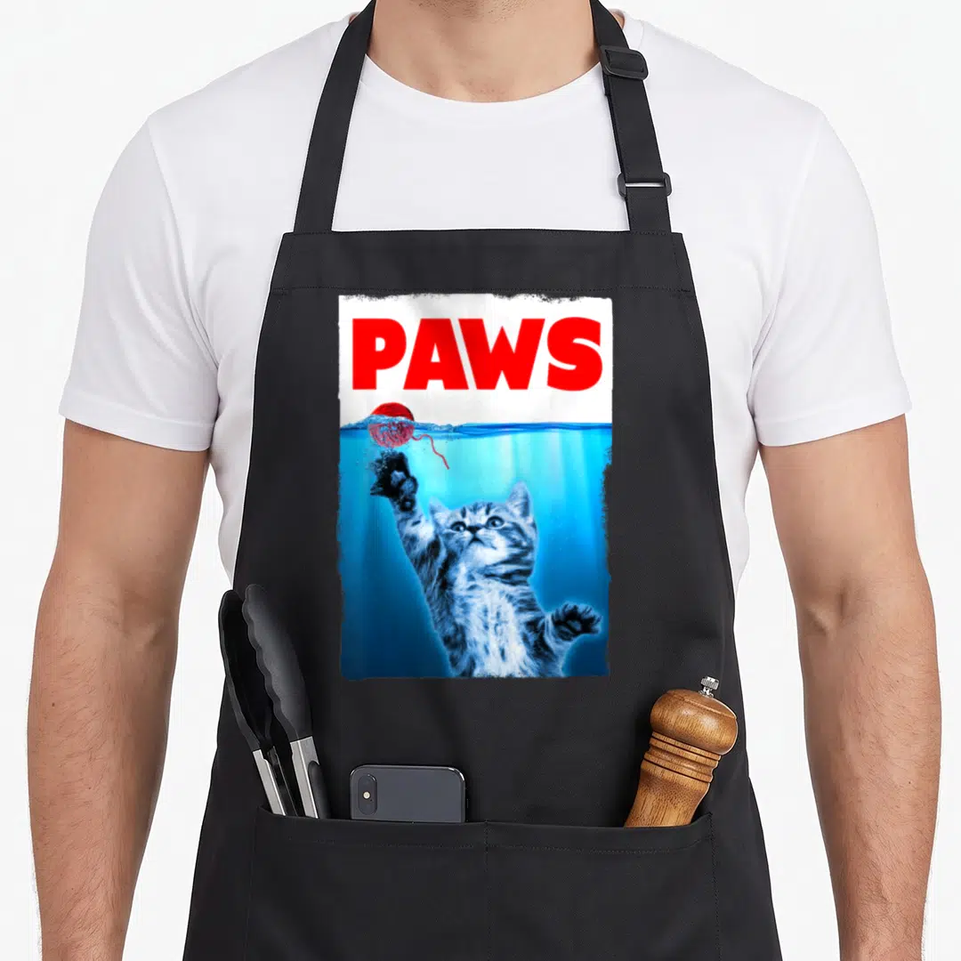 Parody cat apron - PAWS text above underwater kitten reaching for red yarn ball — shown on a black cotton canvas apron against a white background