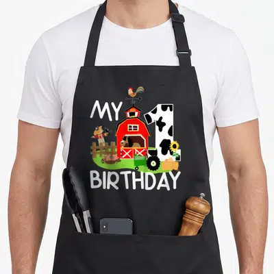 1st chicken apron - Red barn with rooster, cow-print number 1, pig, horse, scarecrow, and MY 1 BIRTHDAY text in cartoon — shown on a black cotton canvas apron against a white background
