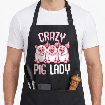 Performance farmer apron - Three cartoon pigs with arched CRAZY text and PIG LADY below in pink and white — shown on a black cotton canvas apron against a white background