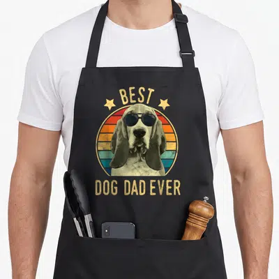 Basset father day apron - Retro dog wearing sunglasses in front of striped sun with BEST DOG DAD EVER text — shown on a black cotton canvas apron against a white background