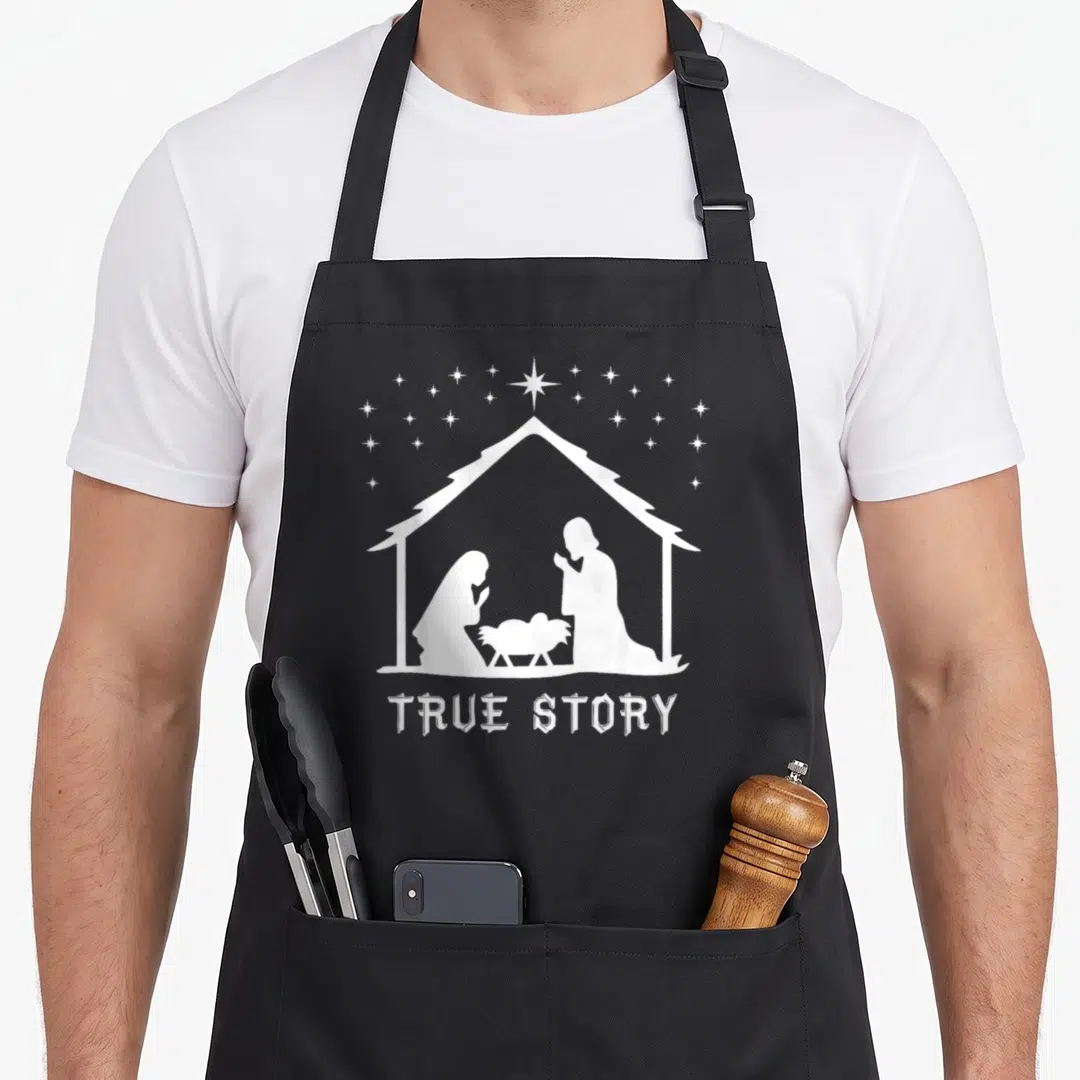 Birth womens christmas apron - White nativity silhouette under stable roof with stars and True Story text — shown on a black cotton canvas apron against a white background