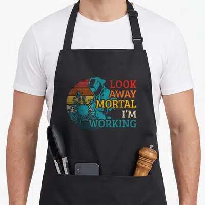 Welding locksmith apron - Retro blue welder silhouette over striped sun with text LOOK AWAY MORTAL I'M WORKING — shown on a black cotton canvas apron against a white background