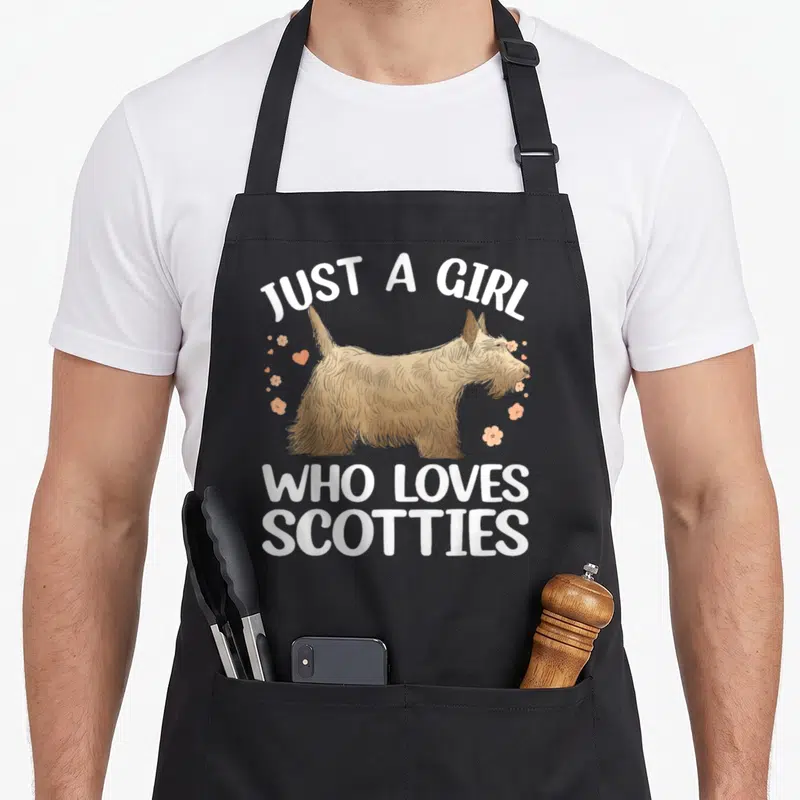 Funny scottish dog apron - Cartoon tan Scottie dog with peach flowers and Just a Girl Who Loves Scotties text — shown on a black cotton canvas apron against a white background