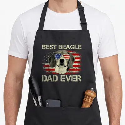 Flag beagle apron - Beagle head wearing sunglasses with a distressed American flag background and beige stencil text — shown on a black cotton canvas apron against a white background