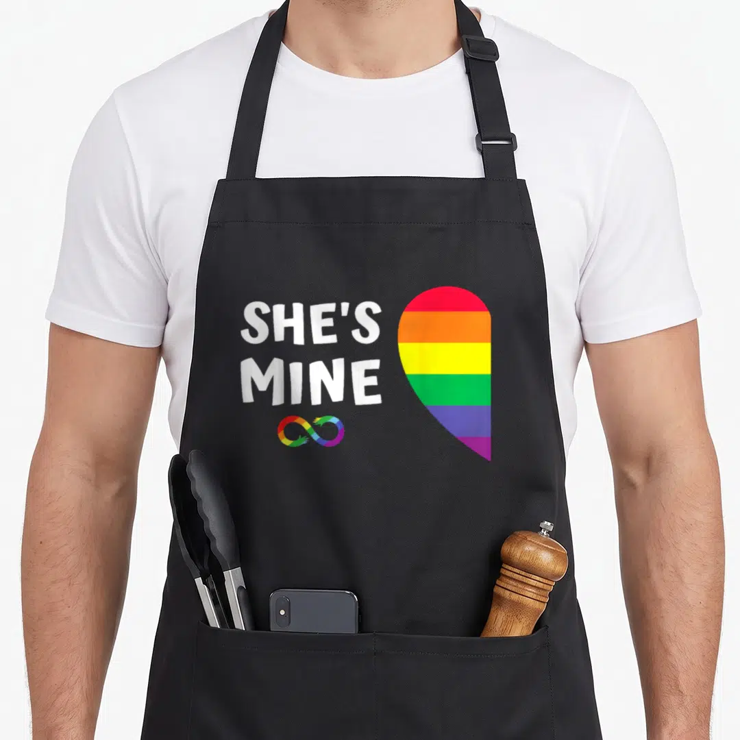 Mine she's in her apron - SHE'S MINE text next to rainbow heart and infinity symbol — shown on a black cotton canvas apron against a white background