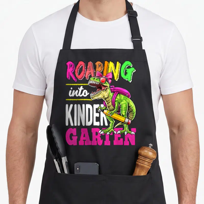 Roaring kindergarten school dinosaur apron - Green T-Rex wearing sunglasses and backpack holding yellow pencil — shown on a black cotton canvas apron against a white background