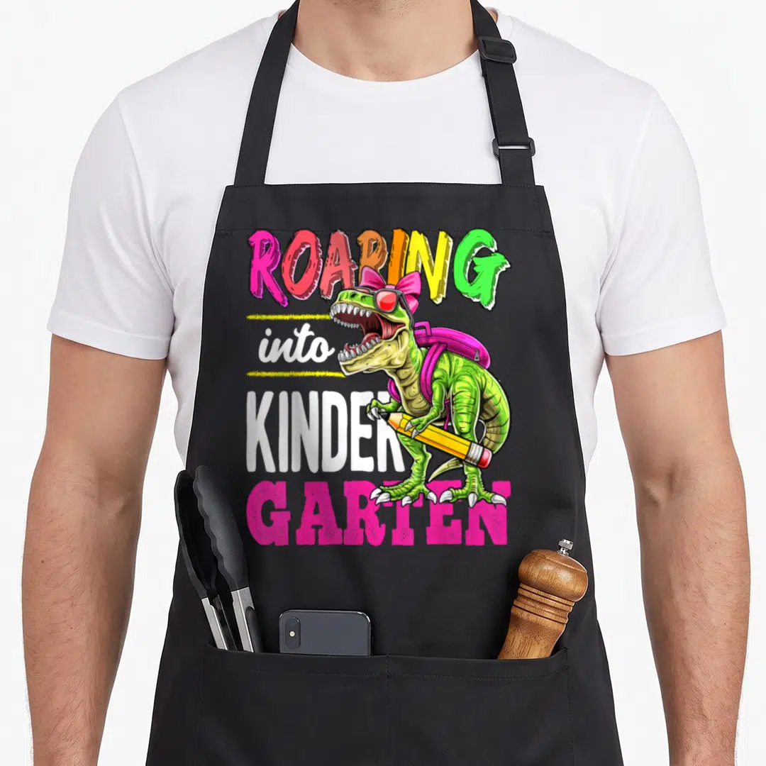 Roaring kindergarten school dinosaur apron - Green T-Rex wearing sunglasses and backpack holding yellow pencil — shown on a black cotton canvas apron against a white background