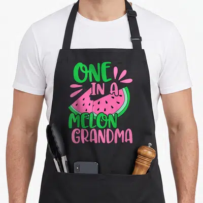 Grandma watermelon apron - Stacked one in a melon grandma text around two cartoon watermelon slices with pink droplets — shown on a black cotton canvas apron against a white background