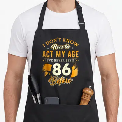 86th birthday apron - Orange and white stacked text reading I Don't Know How To Act My Age 86 Before — shown on a black cotton canvas apron against a white background